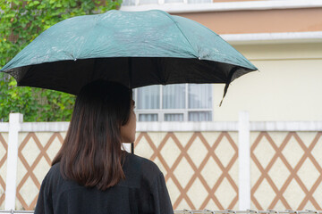 A woman stands under an umbrella in the rain, her back to the camera, gazing at a fence and house.
