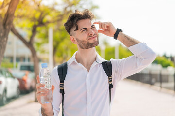Young handsome man with a bottle of water at outdoors having doubts and with confuse face expression