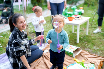 Little girl blows a big soap bubble while standing near her mother sitting on a blanket in the garden