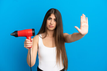 Young caucasian woman holding a hairdryer isolated on blue background making stop gesture