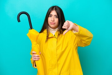 Young caucasian woman with rainproof coat and umbrella isolated on blue background showing thumb...