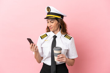 Airplane middle aged pilot woman isolated on pink background holding coffee to take away and a mobile