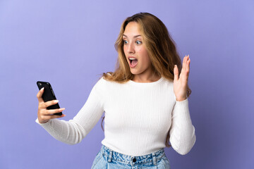 Fototapeta premium Young blonde woman isolated on purple background looking at the camera while using the mobile with surprised expression
