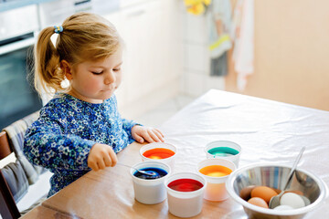Excited little toddler girl coloring eggs for Easter. Cute happy child looking surprised at colorful colored eggs, celebrating holiday with family. Adorable kid at home with different bright colors.