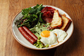 Delicious breakfast plate with fried eggs, grilled sausages, crispy bacon, toasted bread, fresh green salad. Healthy and nutritious morning meal on a wooden table