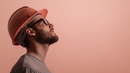 Man wearing a hard hat and glasses. he is standing in front of a plain peach-colored background. the man has a beard and is wearing a grey t-shirt.