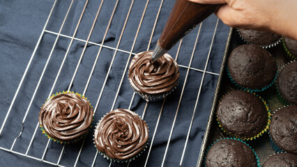 Adding chocolate ganache frosting to chocolate cupcakes on a wire cooling rack.