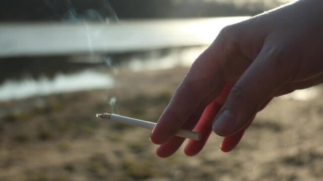 close up of woman hand with lit, smoking cigarette between her fingers against background of spring nature. concept - harm of tobacco, quitting smoking, smoking kill, addiction, abuse, avoid