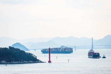 Coastal scene near Busan, South Korea, features multiple sailing ships navigating toward the sea port. A large container ship and a smaller vessel appear in the foreground.