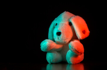 A soft toy in the form of a dog with long ears. The toy is illuminated with multi-colored green and red light. A small red heart is embroidered on one of the ears. The background is dark.