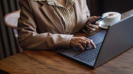 Businesswoman working with laptop on wooden desk, holding smartphone for communication, stressed work and investment decision.