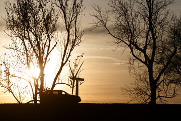 Naklejka premium Sunset silhouette of a vintage car against a scenic landscape with trees and a wind turbine