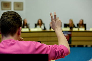 Individual in pink shirt raises hand during discussion at conference with panel of speakers in...