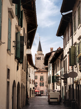 A picturesque narrow street lined with traditional buildings and open shutters leading to a historic bell tower in an Italian town of Venzone with warm sunlight and a relaxed atmosphere