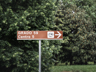 A brown road sign indicating a cycling route on the Alpe Adria Trail near Udine, Italy towards Grado. Lush green trees surround the sign, creating a natural and inviting atmosphere for cyclists.