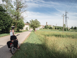 A cyclist pauses on a rural road along the Alpe Adria bike trail near Udine, Italy. Green fields, a small village, and a blue sky create a peaceful summer atmosphere in the countryside.