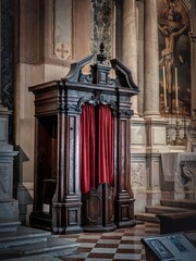 A slightly angled side view of an ornate wooden confessional with a red curtain, highlighting its intricate carvings and timeless religious significance in a historic church interior.