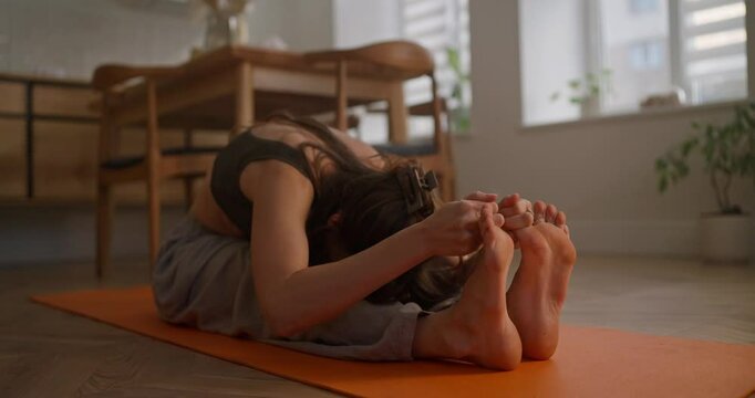 Focused brunette girl doing stretching and sports and reaching her head to her feet while sitting on an orange mat at home