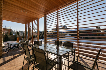 This outdoor dining space showcases a sleek table surrounded by chairs, enhanced by slatted wood panels. Snow covers the ground, creating a serene winter atmosphere.