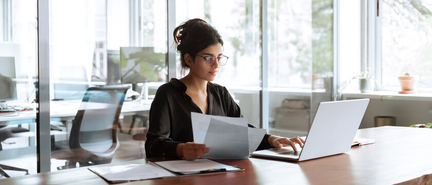 Businesswoman holding document working at laptop pc computer doing online tax invoice, financial report. Focused Hispanic business woman manager specialist with paperwork in office. Banner, copy space