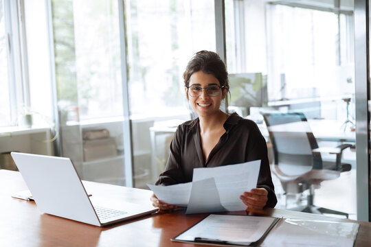 Smiling portrait at camera business woman accountant holding documents, working at laptop pc computer doing online tax invoice, financial report. Latina businesswoman manager with paperwork in office 