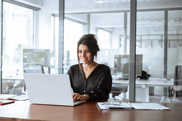 Smiling young latina hispanic business woman working on laptop computer in office. Focused Indian businesswoman professional manager IT specialist using AI pc app, thinking, make analysing. Copy space