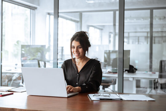 Smiling young latina hispanic business woman working on laptop computer in office. Focused Indian businesswoman professional manager IT specialist using AI pc app, thinking, make analysing. Copy space