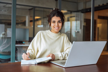 Portrait young latina or indian business woman using laptop pc working at desk writing notes,...