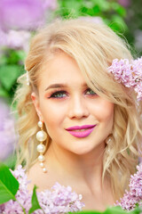Fototapeta premium Blonde with blue eyes and pink lipstick stands with purple flowers. She is wearing pearl earrings and a pink dress. The image has a soft and romantic mood, with the beauty of the woman. Close-up portr