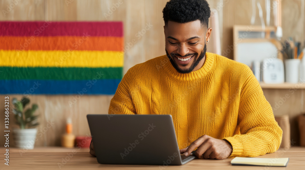Poster man in yellow sweater smiles while using laptop at wooden desk, with colorful pride flag in backgrou