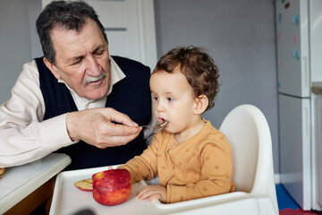 A grandfather joyfully feeds his grandson in a cheerful kitchen, creating fond memories during a...