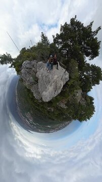 Vertical video. A Couple Sitting on a Rocky Peak in a Tiny Planet 360-Degree Spherical Panorama Surrounded by Sky and Forest with a Dramatic Cloudy Backdrop