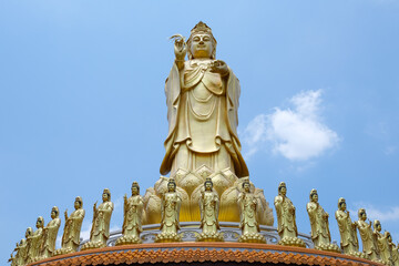 A magnificent golden Guanyin statue standing tall against a clear blue sky, surrounded by smaller...