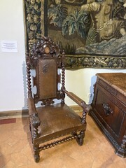 Wooden ornate chair with intricate carvings placed against a tapestry-covered wall in a museum...