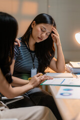 Stressed Asian businesswoman having headache at office desk, receiving support from colleague