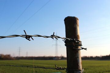 Close-up of a barbed wire fence and post with green meadow, power transmission and blue sky in the background