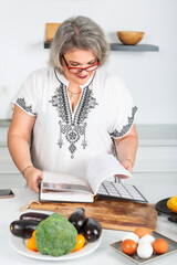 Beautiful senior woman looking at recipe book while cooking in kitchen. Healthy eating. Weight loss concept. Vitamin foods are good for your health. Blogger. Housewife.	