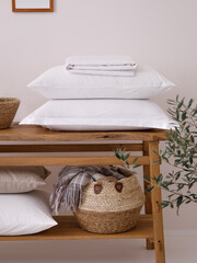 Rustic oak shelves with home textiles, including a woolen blanket placed in a basket, four pillows and a white sheet in the modern bright bathroom. Close-up photo. Front view.