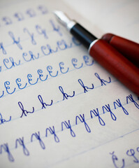Close-up of uppercase and lowercase letters written by a child in cursive with a fountain pen with blue ink in a lined notebook, a burgundy fountain pen in the background.