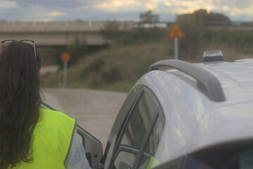 Woman with her back turned wearing a vest with the V16 emergency beacon on the roof of the broken-down vehicle. The emergency flasher is mandatory for replacing triangles in Europe. DGT Spain.