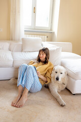 Relaxed woman in a warm sweater and striped pajama pants lounges on a soft carpet, cuddling her...