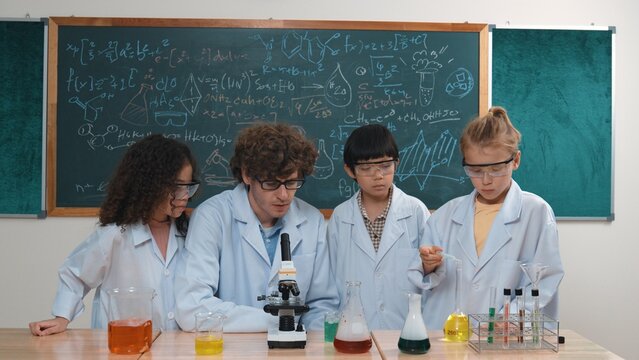 Smart caucasian teacher looking under microscope while diverse children doing experiment at STEM science lesson. Professional happy scientist inspect biochemical example by using microscope. Pedagogy.