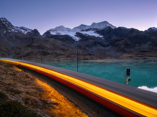 Moving modern red train near azure mountain Bianco lake, snowy rocks and purple sky at night in...