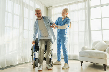 Rehabilitation healthcare. Physiotherapist nurse helps old man patient in wheelchair stand up...