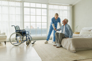 Rehabilitation healthcare. Physiotherapist nurse helps old man patient in wheelchair stand up...