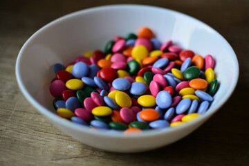 A full bowl of colorful candies close up view on wooden table. Chocolate buttons sweet treats in white bowl.
