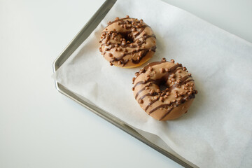 Two brown cappuccino donuts on paper, metal tray and white table