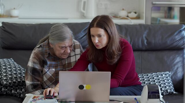 In a cozy home setting, a daughter guides her senior mother in using a laptop, helping her learn essential tech skills.