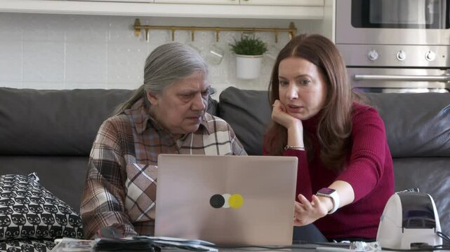 A middle-aged woman patiently instructs her elderly mother on how to use a laptop for online store trading while sitting together on a sofa at home.