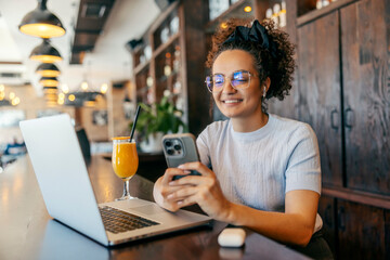 Portrait of young woman sitting in cafe with cellphone in hands. There is a laptop on table.